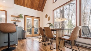 A modern kitchen with wooden flooring, a dining area with chairs, and a large window offering a view of trees. - Blue Horizon by Taylor Made Deep Creek (Swanton)