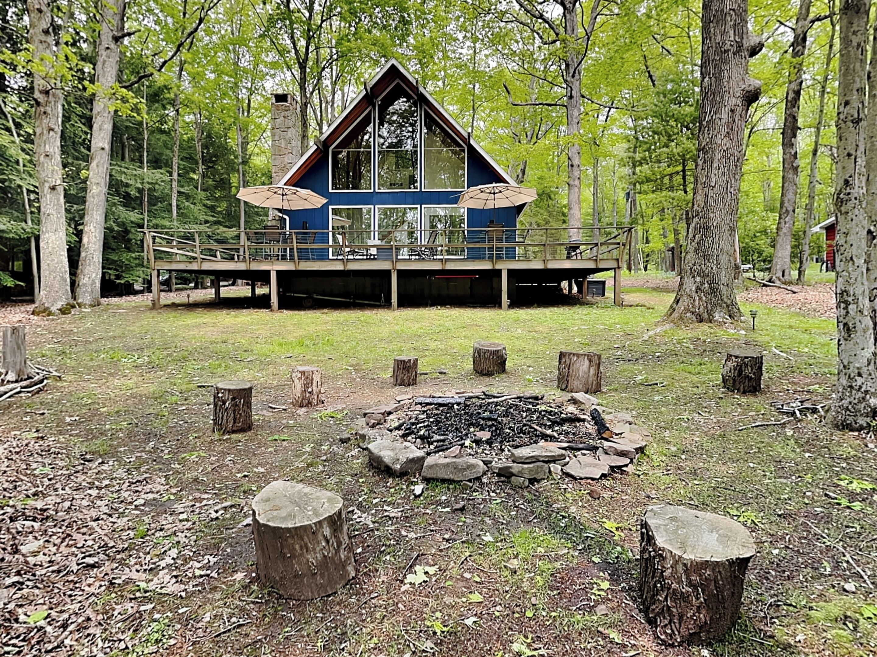 A cabin with a deck, surrounded by trees and a fire pit.