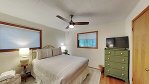 A bedroom with a bed, a ceiling fan, a TV, a dresser, a lamp, a window with blinds, and a small table with a basket. - Blue Horizon by Taylor Made Deep Creek (Swanton)