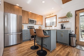A modern kitchen with a stainless steel refrigerator, a microwave, and a coffee maker. There are two bar stools and a sink with a faucet.