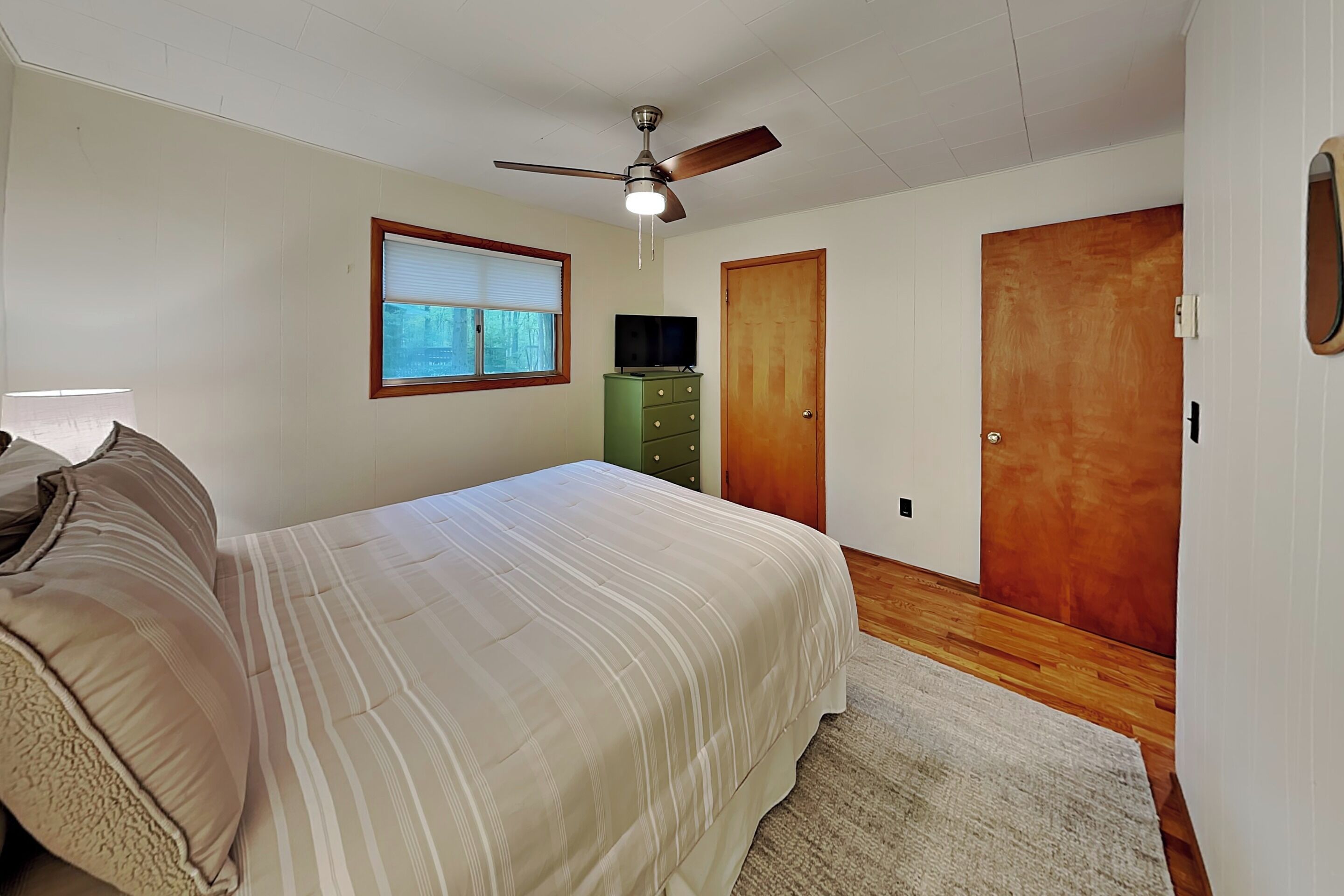 Un dormitorio con cama, ventilador de techo, ventana, cómoda y dos puertas.