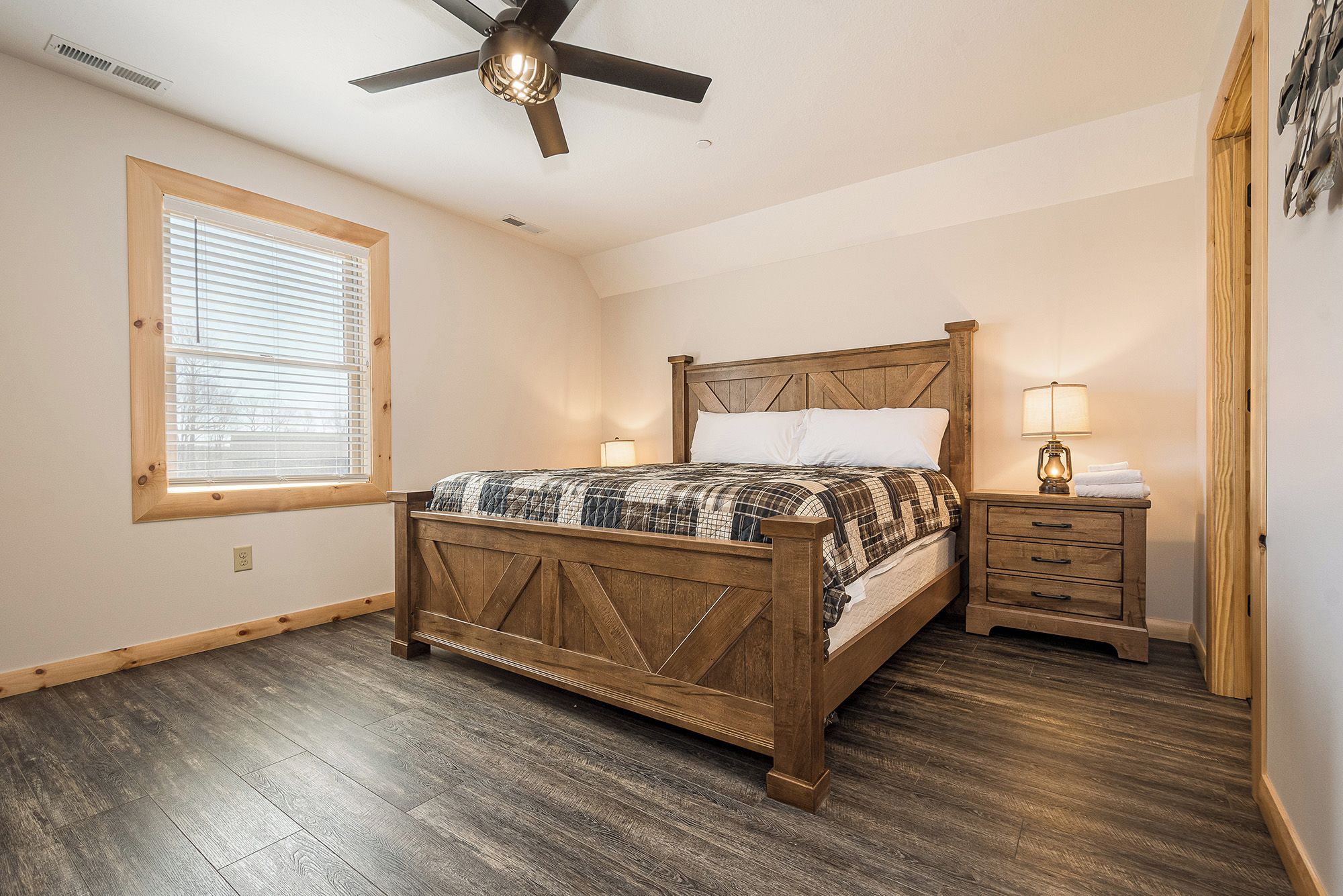 A bedroom with a wooden bed, a nightstand, a ceiling fan, a window with blinds, and a wooden floor.