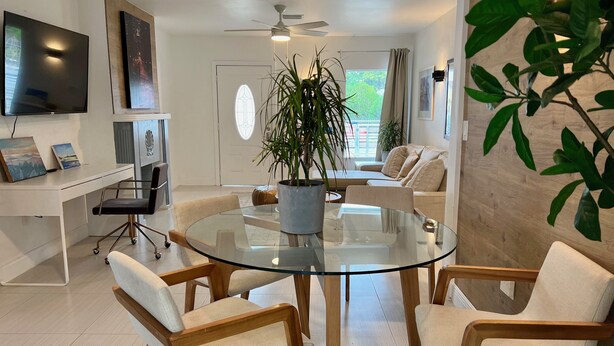 A modern living room with a glass-top coffee table surrounded by four chairs, a white sofa, a TV mounted on the wall, and a ceiling fan. - Centrally located 5 bedroom house with parking (Miami)
