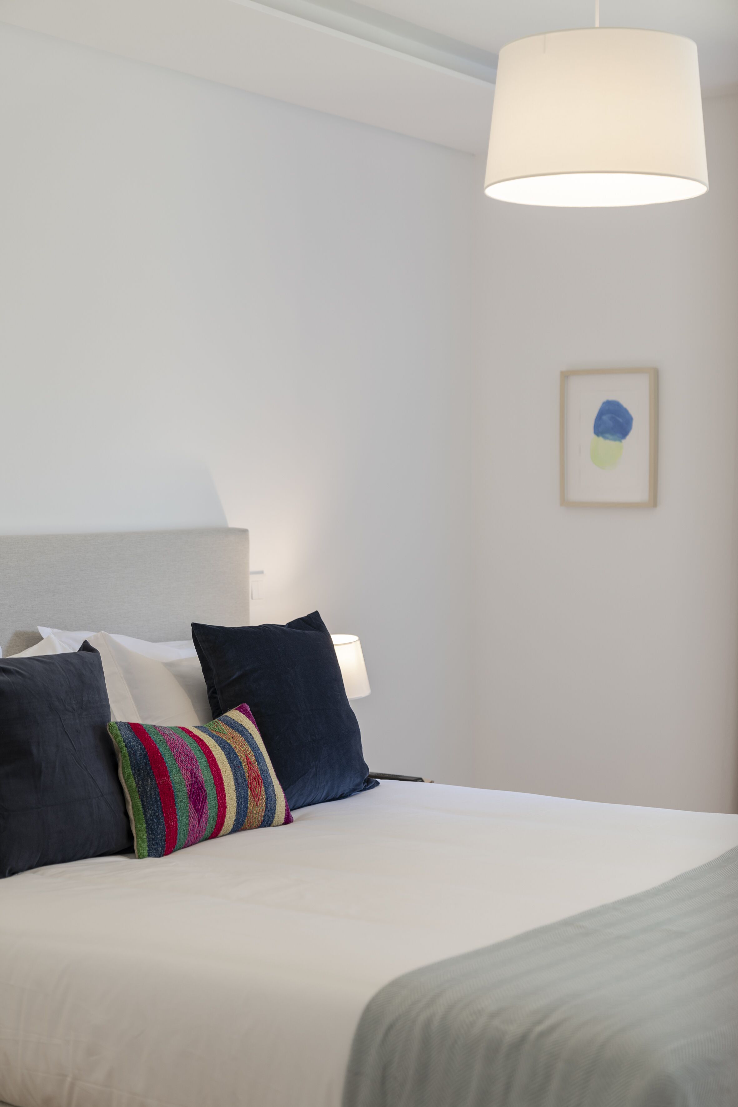 A neatly made bed with a white comforter and multiple pillows, including a colorful striped one. There is a white lamp hanging above the bed and a framed abstract painting on the wall.