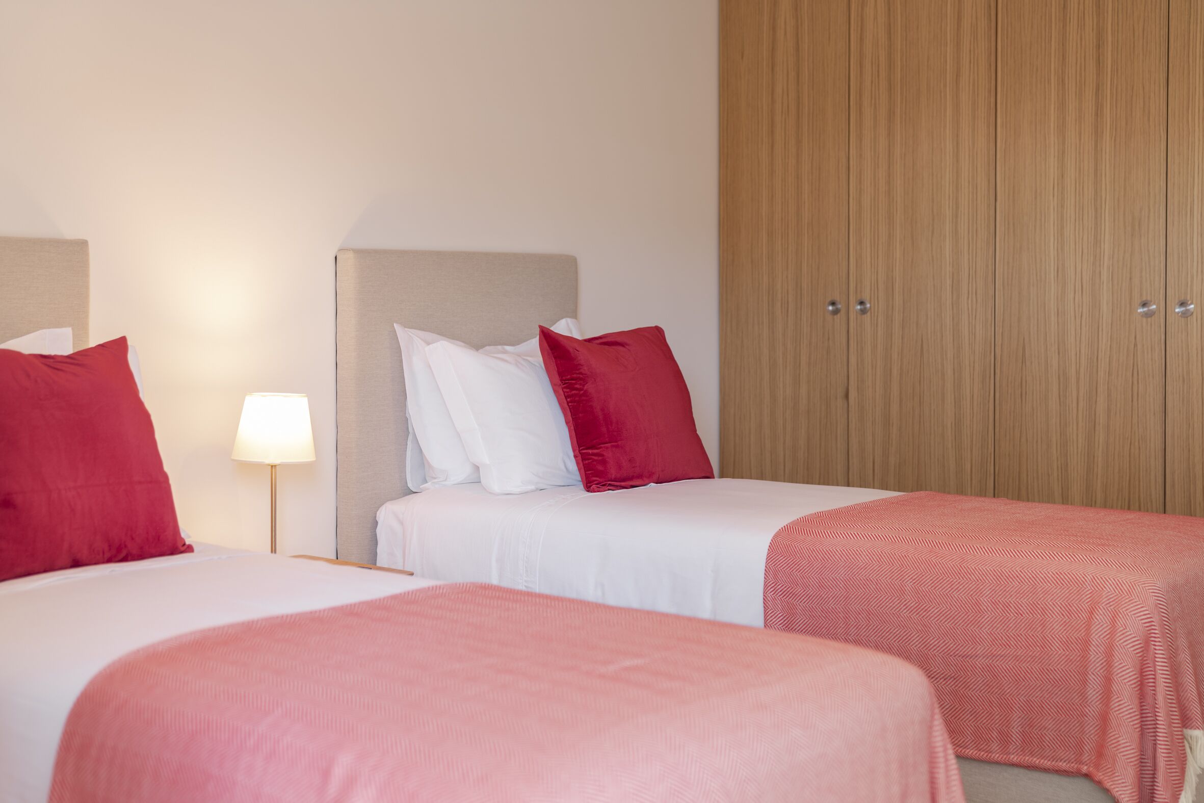 Habitación de hotel con dos camas, cada una con ropa de cama roja y blanca, una mesita de noche con lámpara y un armario de madera.