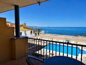 A balcony with a view of the sea, a pool, and a building with a chimney.
