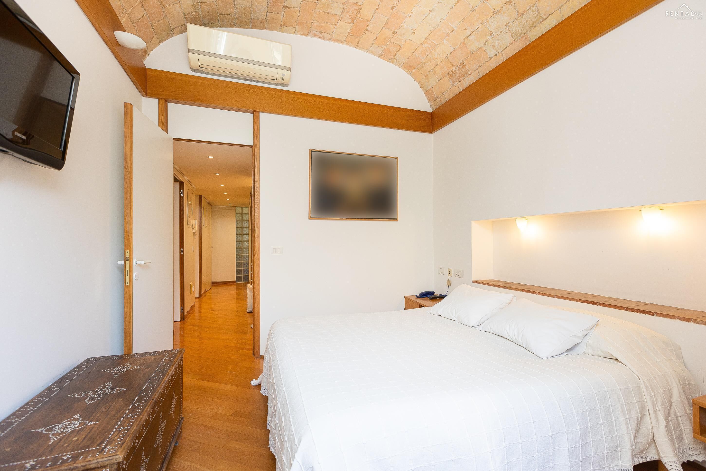 A bedroom with a large bed, a wooden headboard, a mounted TV, and a ceiling fan.