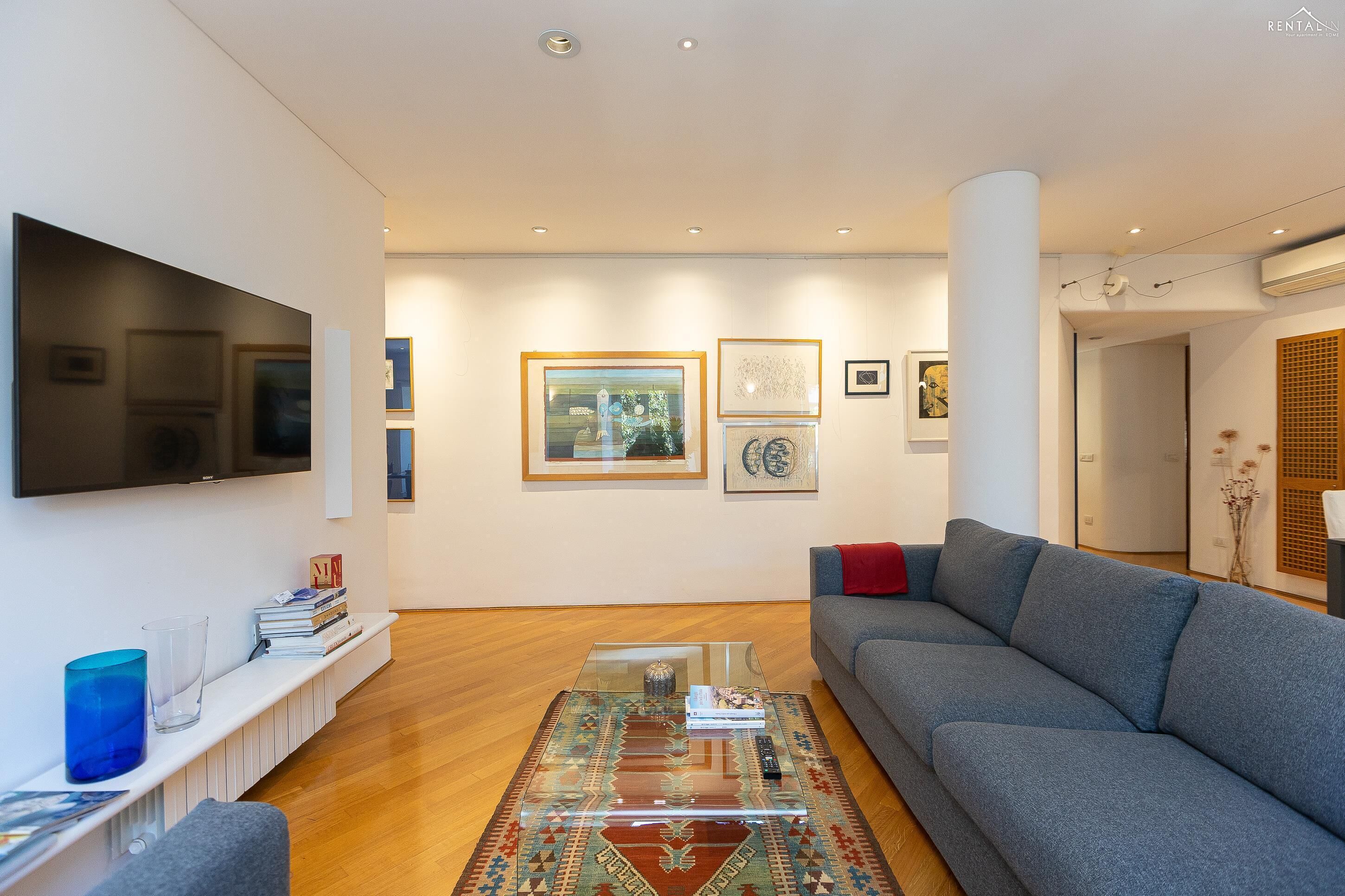 A modern living room with a grey sofa, a glass coffee table, and a flat-screen TV mounted on the wall.
