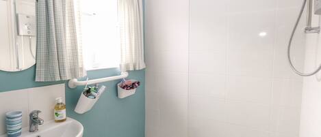 A bathroom with a sink, a mirror, a window with curtains, a shower, and toiletries on the wall.