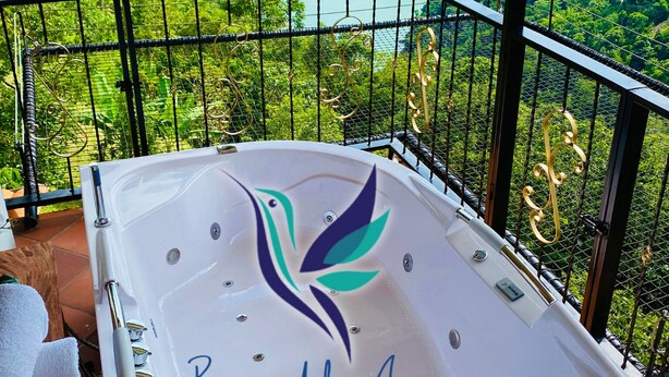 Een bubbelbad met een logo en de tekst 'Brisas del Amanecer Ecohotel' op een balkon met uitzicht op een weelderige vallei en rivier.