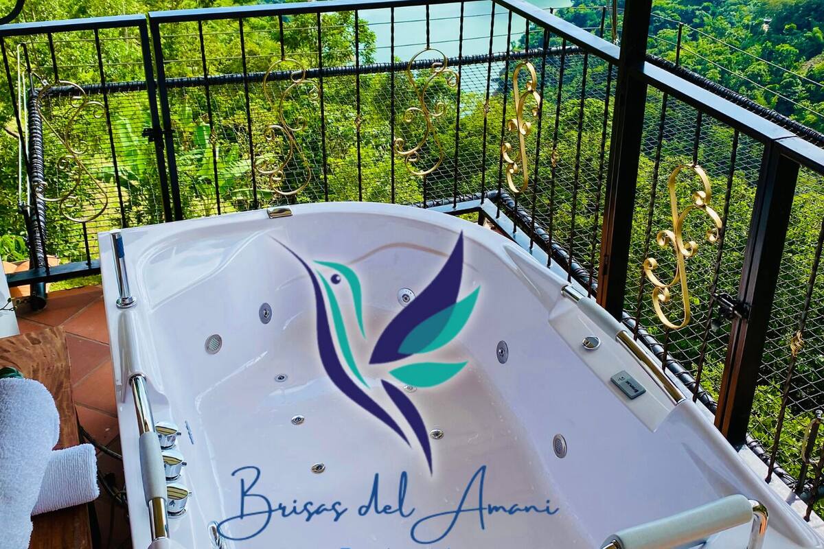 Un bain à remous orné d’un logo et de l’inscription « Brisas del Amanecer Ecohotel », situé sur un balcon donnant sur une vallée verdoyante et une rivière.