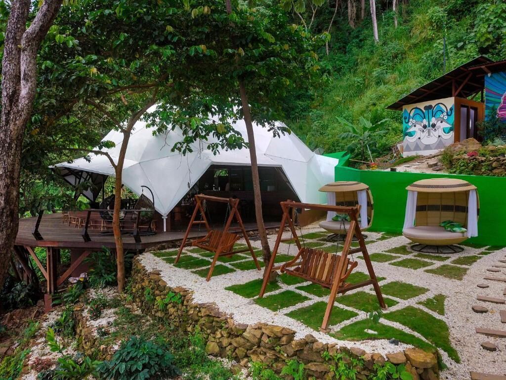 Una estructura tipo tienda blanca con un suelo de madera y columpios, rodeada de vegetación y un muro de piedra.
