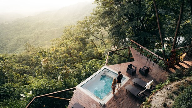Una terraza de madera con piscina, rodeada de exuberante vegetación y con vistas a un valle boscoso.