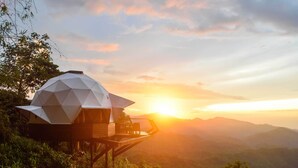 Estructura de domo geodésico con terraza y zona de estar, con montañas al fondo durante el atardecer.