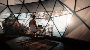Una persona sentada en un escritorio dentro de una estructura en forma de domo geodésico, con una computadora portátil, ventanas amplias y vistas a árboles y montañas.