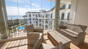 A balcony with wicker furniture, a glass balustrade, and a view of a pool and buildings. - Marina de la Duquesa 544 (Manilva)