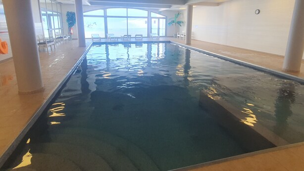 Une piscine intérieure à l’eau claire, entourée de murs blancs et de grandes fenêtres.