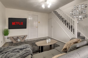 A living room with a flat-screen TV displaying "NETFLIX," a staircase, and a wall of framed pictures.