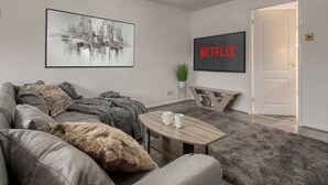 A modern living room with a grey sofa, a coffee table with two mugs, a framed cityscape painting, and a 'NETFLIX' sign on the wall.