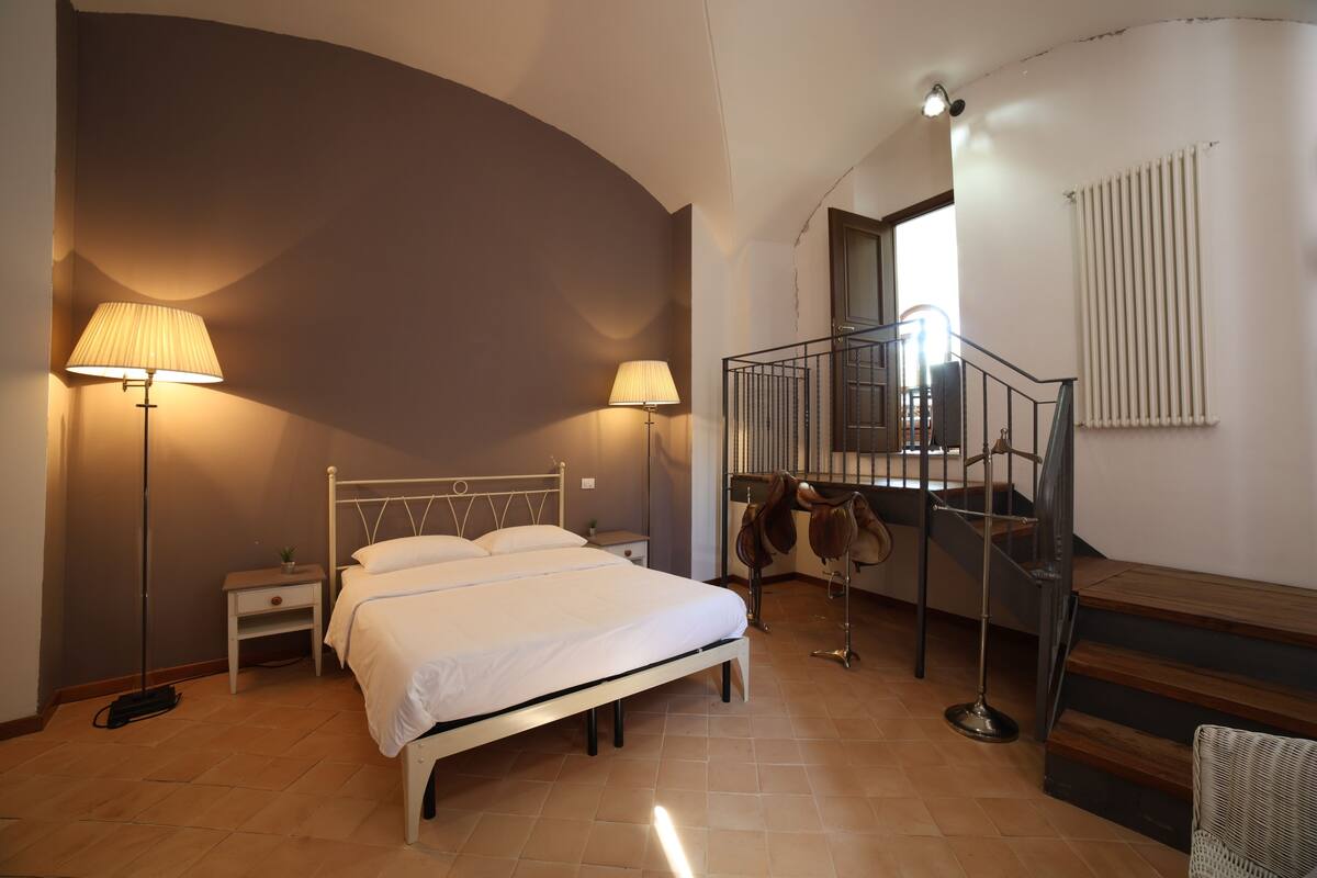 A bedroom with a bed, two bedside lamps, a nightstand, a radiator, and a staircase with a railing.