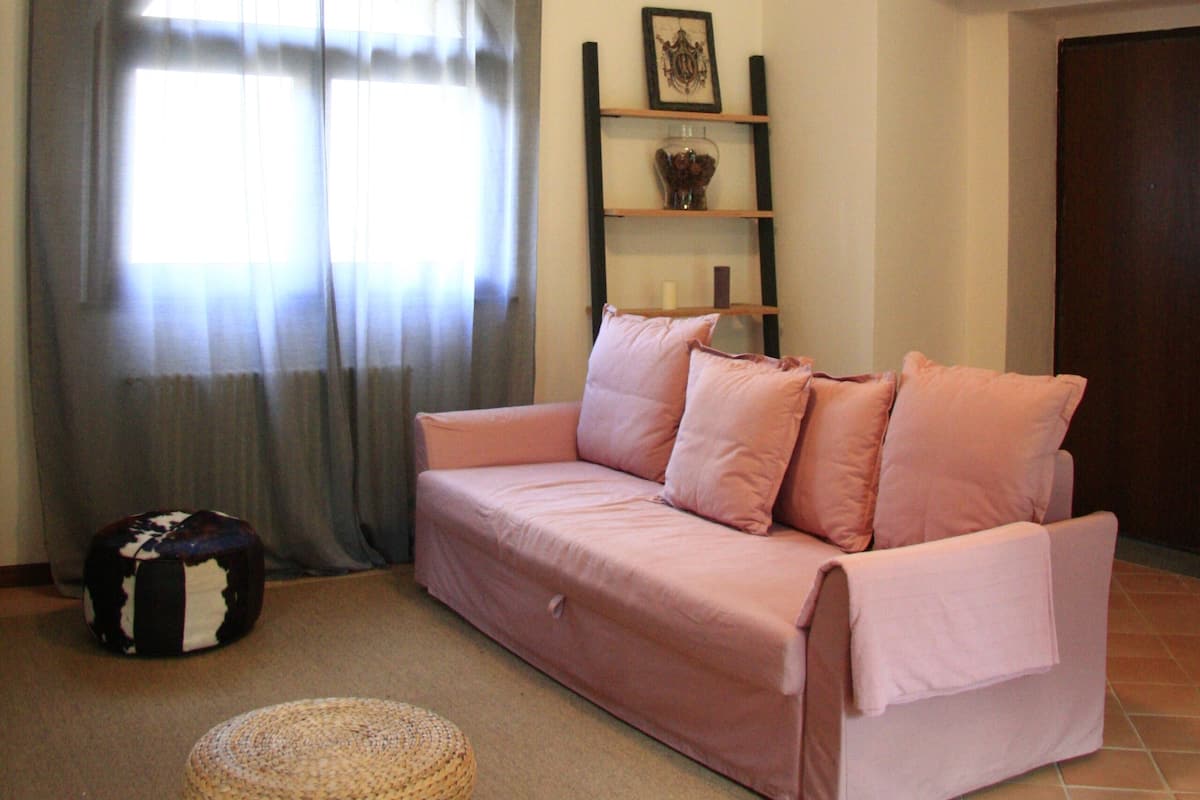 A living room with a pink sofa, a round woven ottoman, a white table, a bookshelf with decorative items, and a window with curtains.