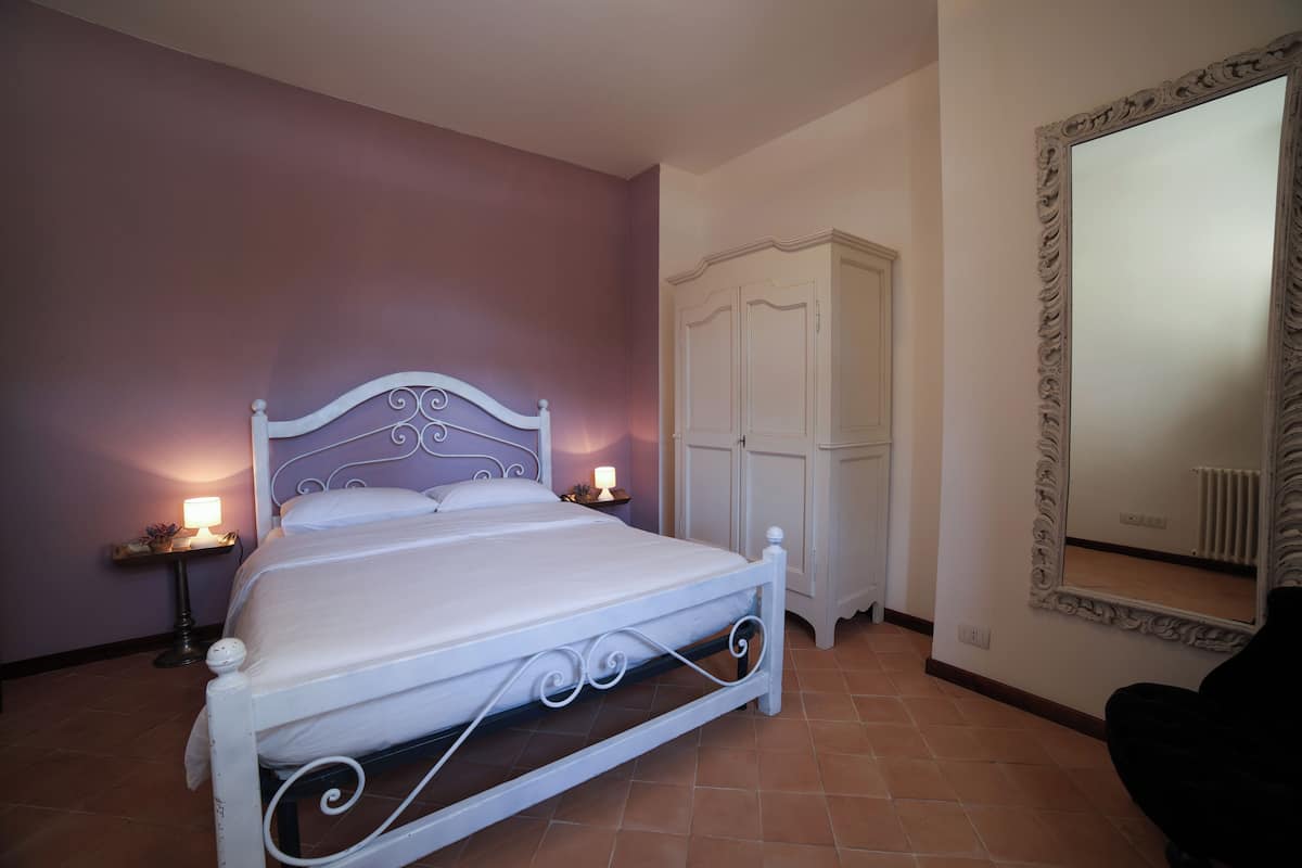 A neatly made bed with a white headboard and footboard, a bedside table with a lamp, a wardrobe, and a large mirror on the wall.