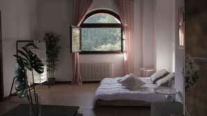 A bedroom with a bed, bedside table, window, radiator, curtains, and a potted plant. - HT CASTELLO DI CAMPOLONGO (Valfabbrica)