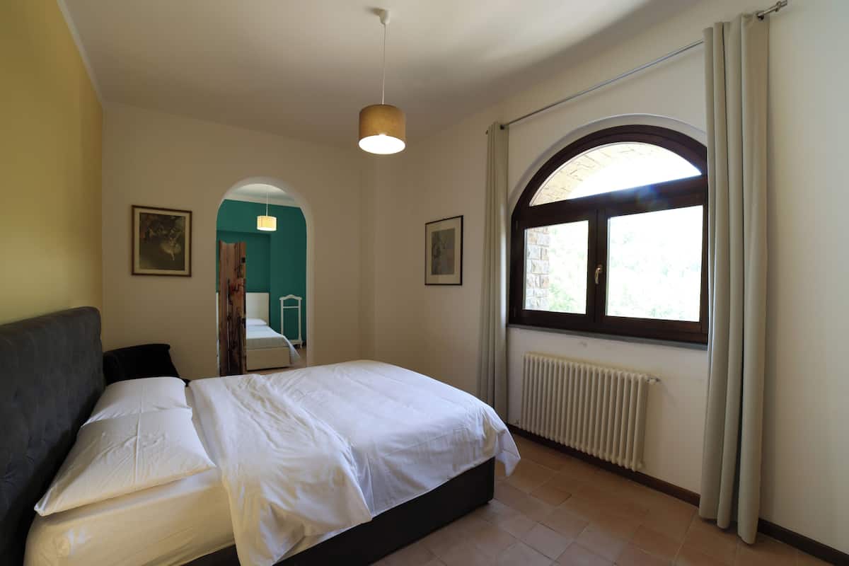A bedroom with a bed, a radiator, a window with curtains, and a door leading to another room.