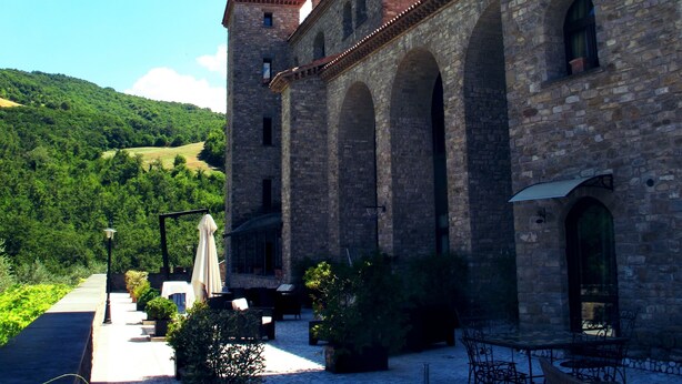 A courtyard with stone flooring, outdoor seating, and a stone building with arches and a tiled roof. - HT CASTELLO DI CAMPOLONGO (Valfabbrica)