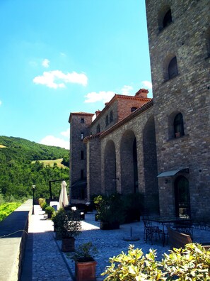 A courtyard with stone flooring, outdoor seating, and a stone building with arches and a tiled roof. - HT CASTELLO DI CAMPOLONGO (Valfabbrica)
