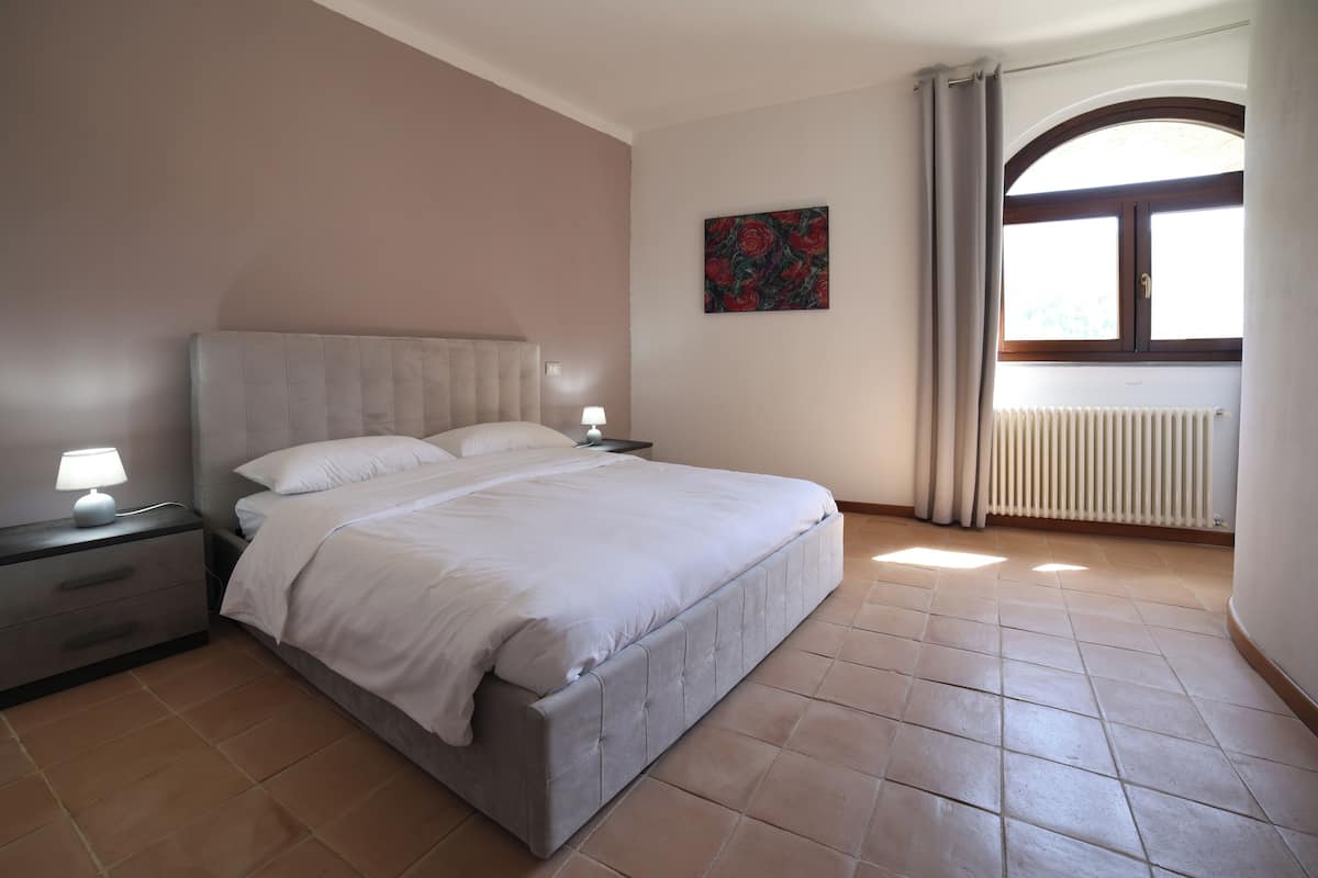 A bedroom with a large bed, a nightstand, a lamp, a radiator, and a window with curtains.