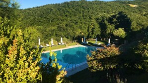A pool with lounge chairs and umbrellas, surrounded by lush greenery and hills. - HT CASTELLO DI CAMPOLONGO (Valfabbrica)