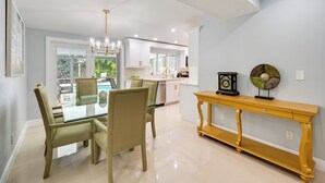 A modern dining area with a glass table and chairs, a yellow console table, and a view of the kitchen and outdoor area. - Pelican Nest, Tropical Naples Villa w/ Pool (Naples)