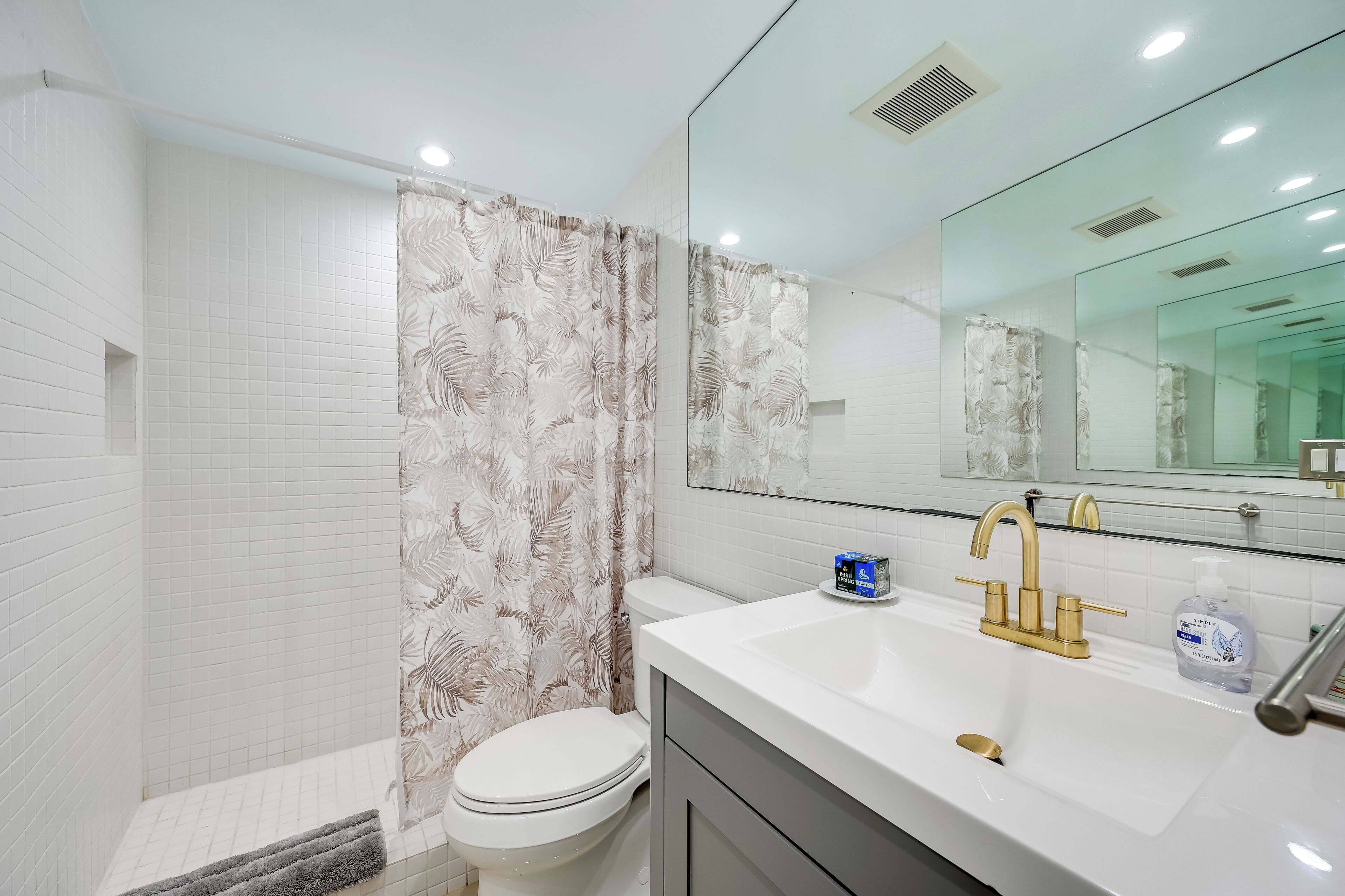 A modern bathroom with a white tiled shower, a toilet, a sink with a gold faucet, and a large mirror.