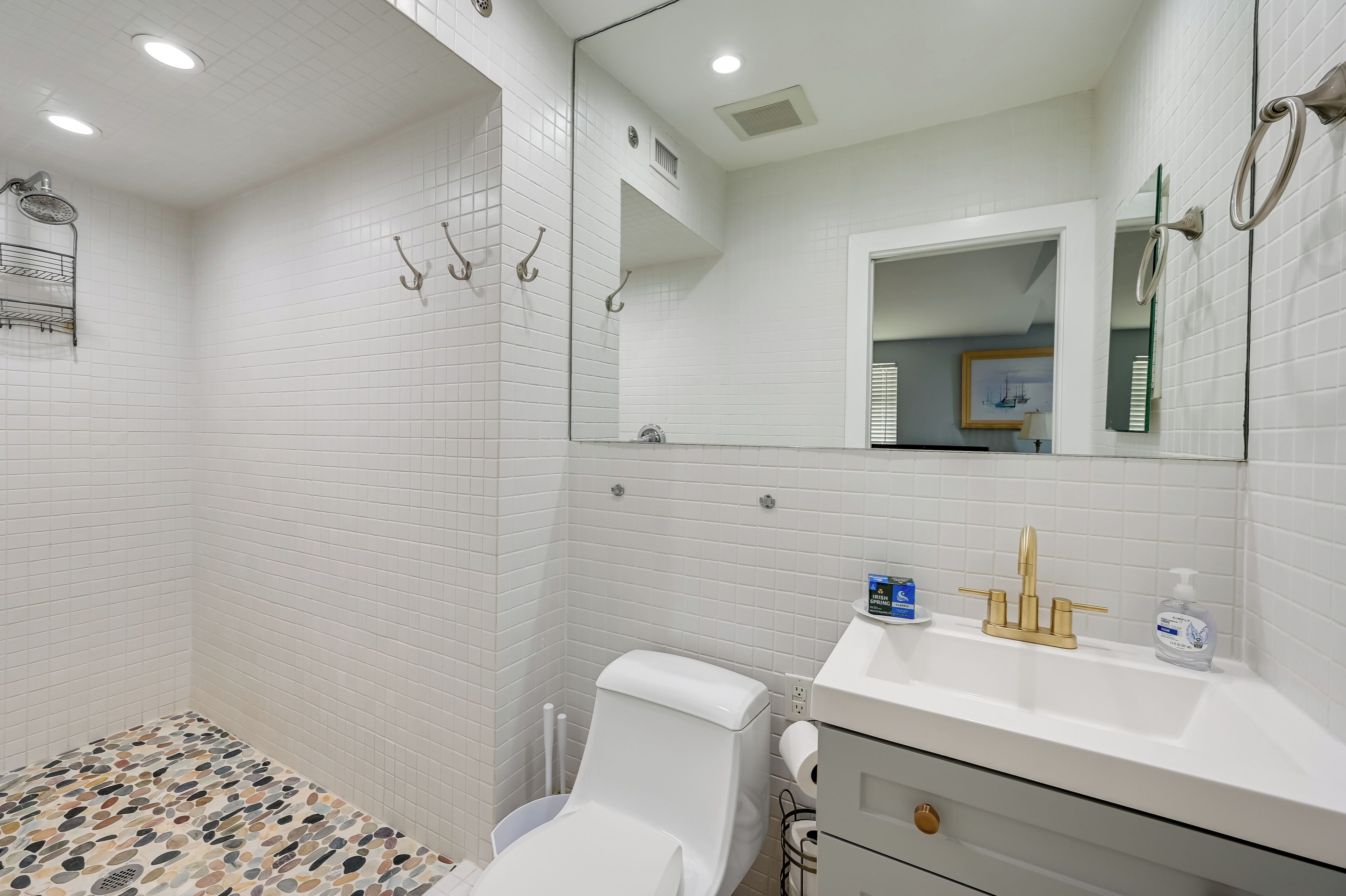 Een badkamer met witte betegelde wanden, een douche, toilet en een wastafel met gouden kraan, en een spiegel.
