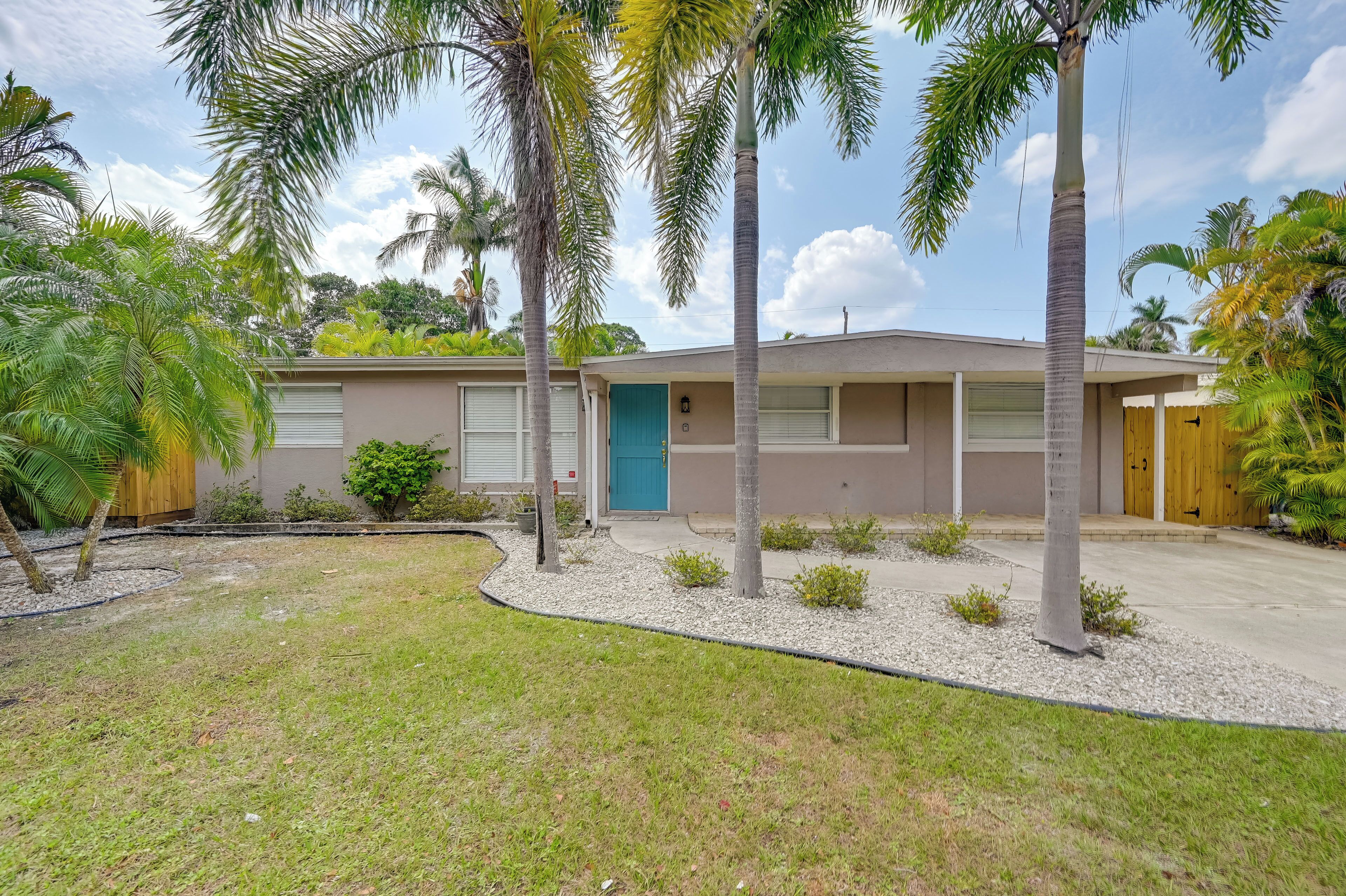 A single-story house with a front garage, a blue door, and a small front yard with palm trees and a gravel pathway.
