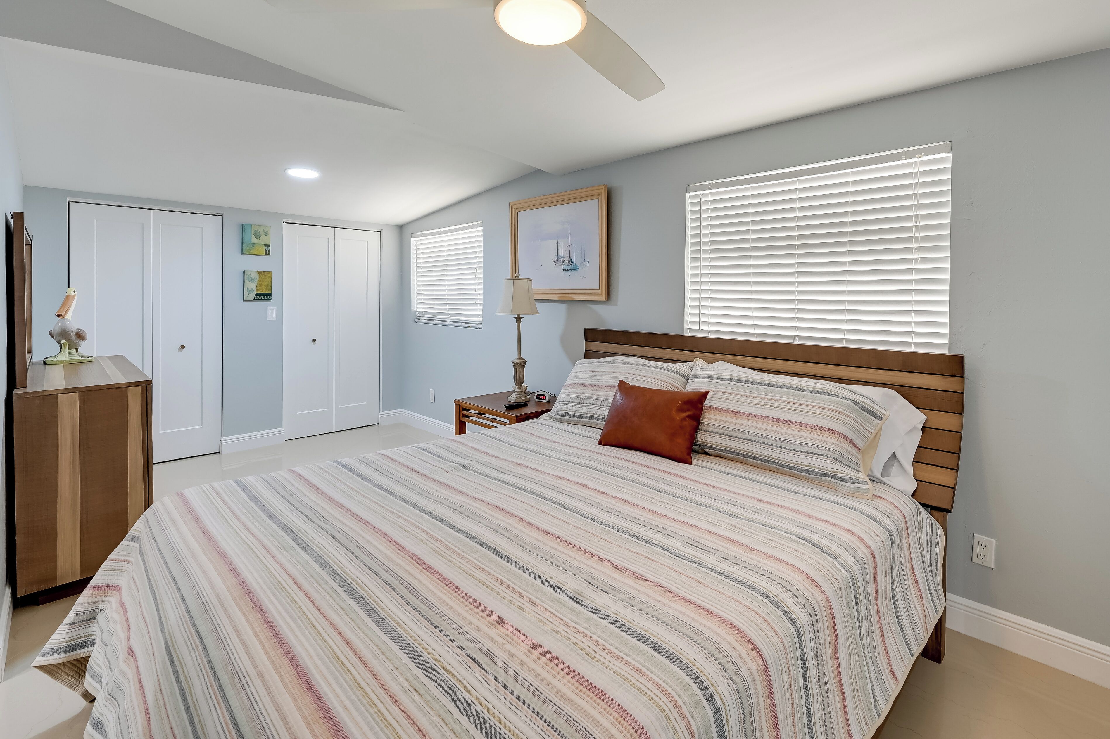 Un dormitorio con una cama grande, cabecera de madera, mesita de noche, lámpara y ventilador de techo. Hay dos puertas y una ventana con persianas.