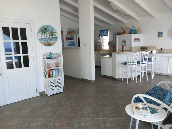 Un'ampia cucina con mobili bianchi, un'area pranzo con tavolo e sedie bianche, e vista sul mare tramite una porta.
