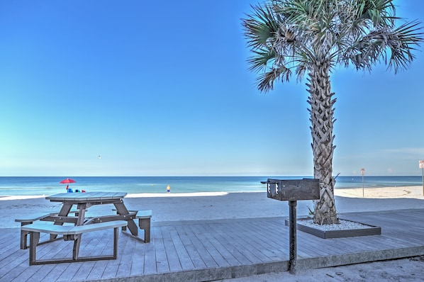 A beach with a wooden deck, picnic tables, a palm tree, and a clear blue sky.