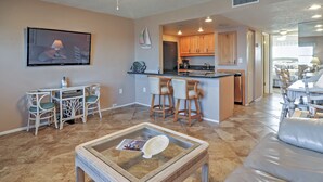 A modern kitchen with wooden cabinets, a central island, and a dining area with white chairs. - Beachfront St Petersburg Condo w/ Community Pool! (Treasure Island)