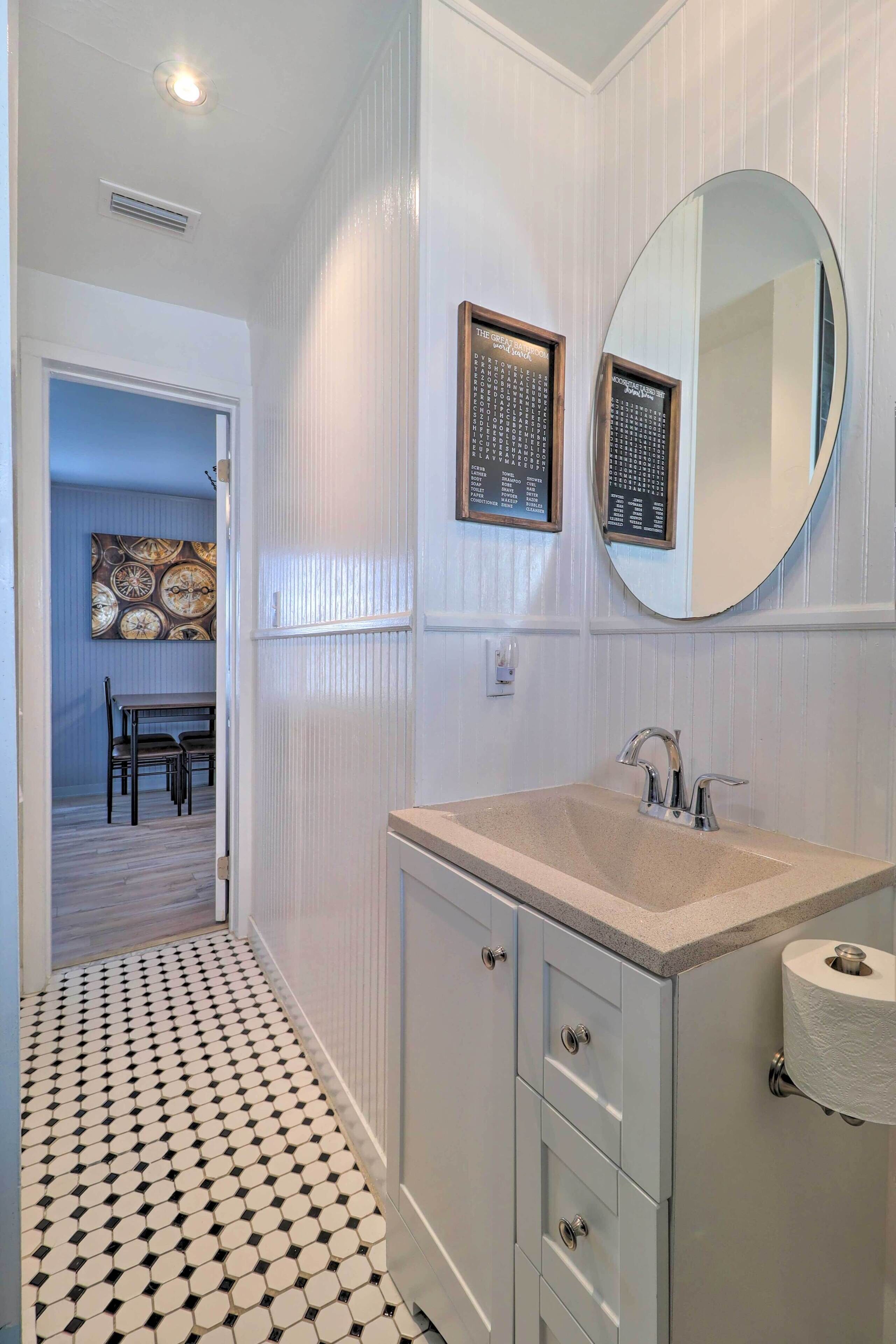 A bathroom with a sink, mirror, and toilet paper holder. There is a door leading to a dining area with a table and chairs.