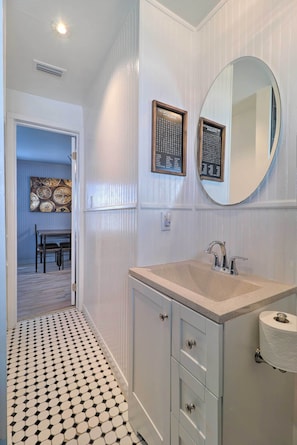 A bathroom with a sink, mirror, and toilet paper holder. There is a door leading to a dining area with a table and chairs.