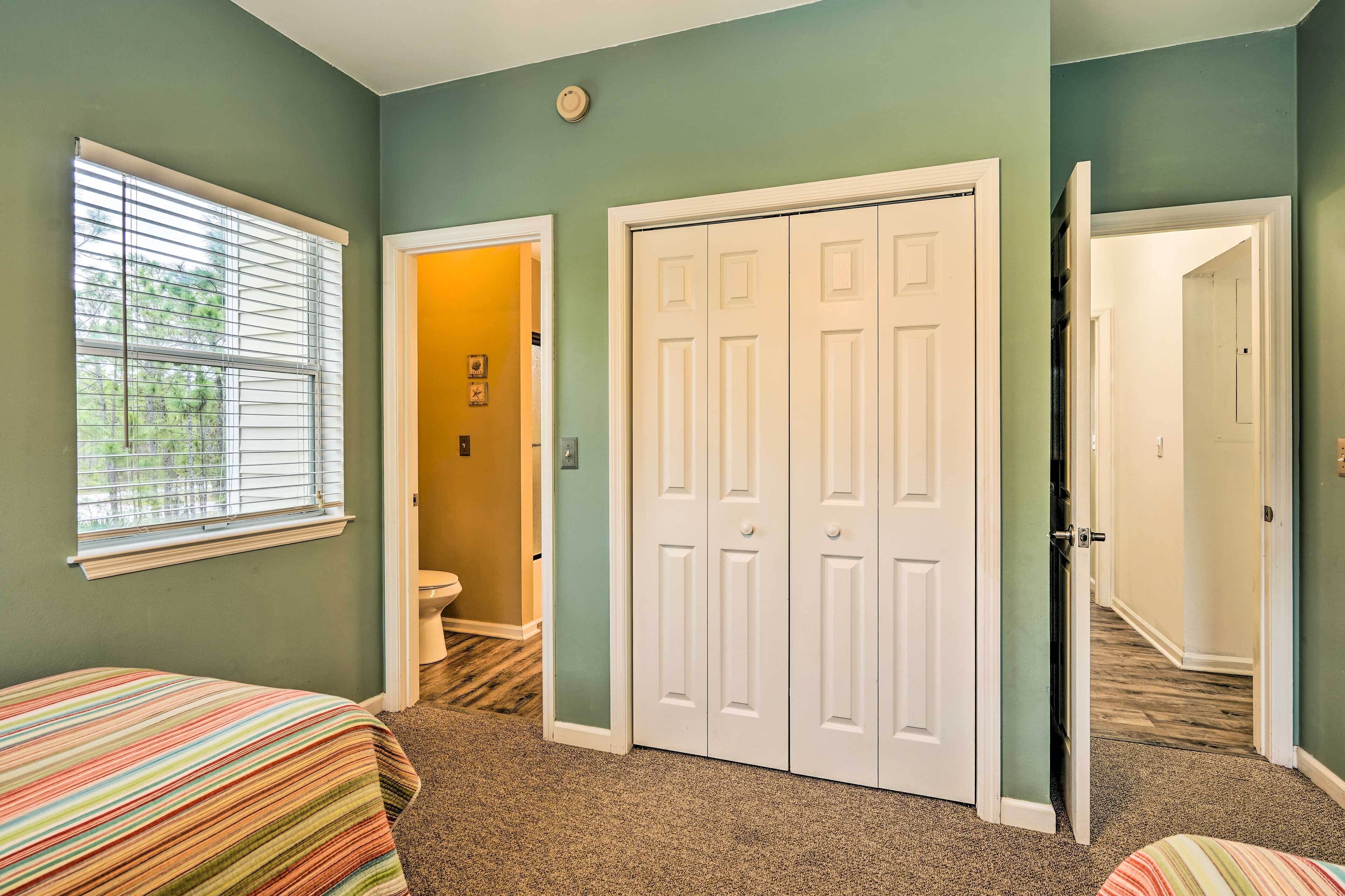 Une chambre avec deux lits, une fenêtre avec des stores, un placard à deux portes et une salle de bain visible à travers une porte ouverte.