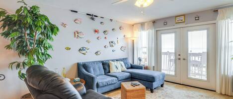 Un salon avec un canapé bleu, un fauteuil en cuir noir, une grande plante en pot et un ventilateur de plafond.