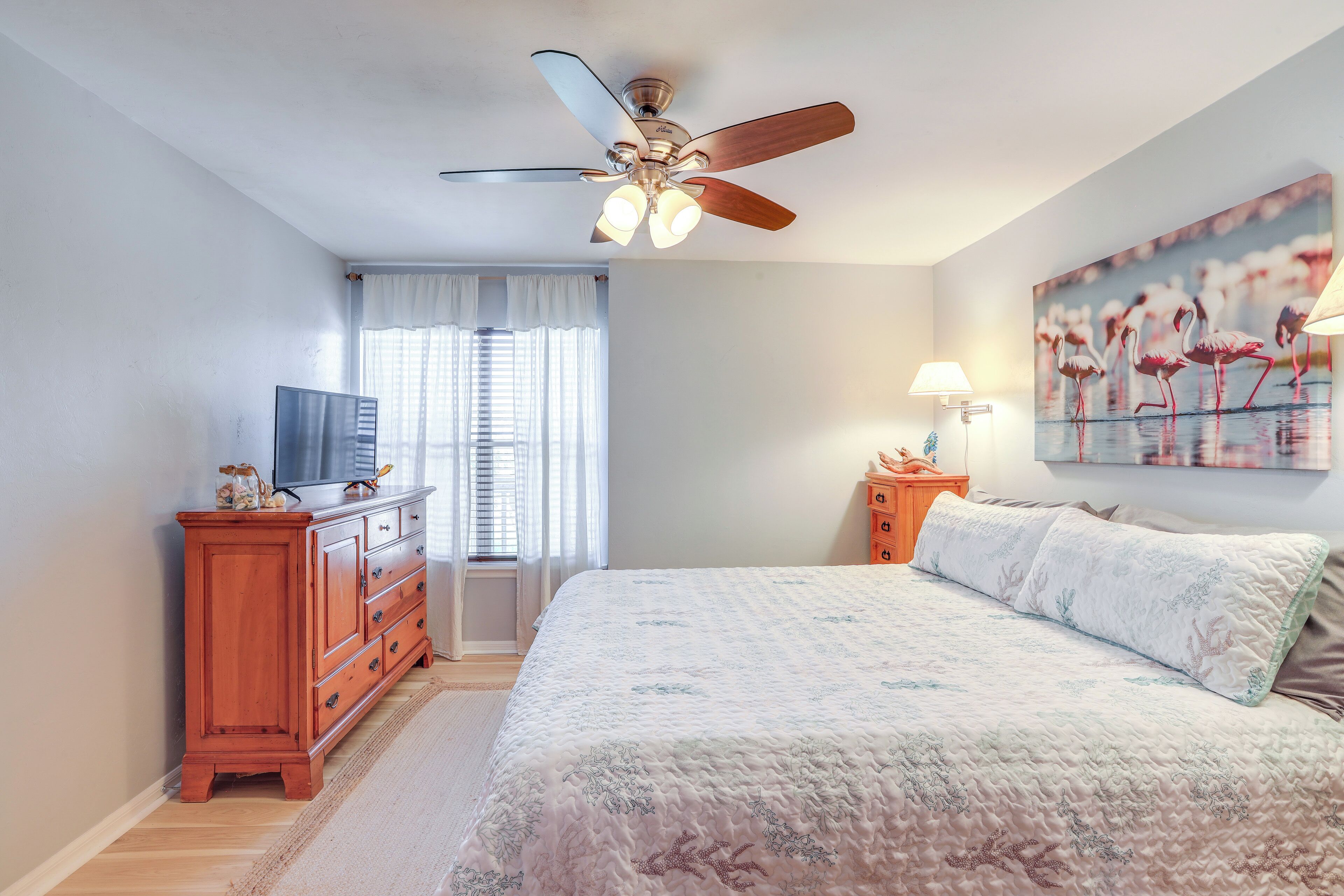 A bedroom with a bed, a ceiling fan, a TV, a dresser, a lamp, and a window with curtains.