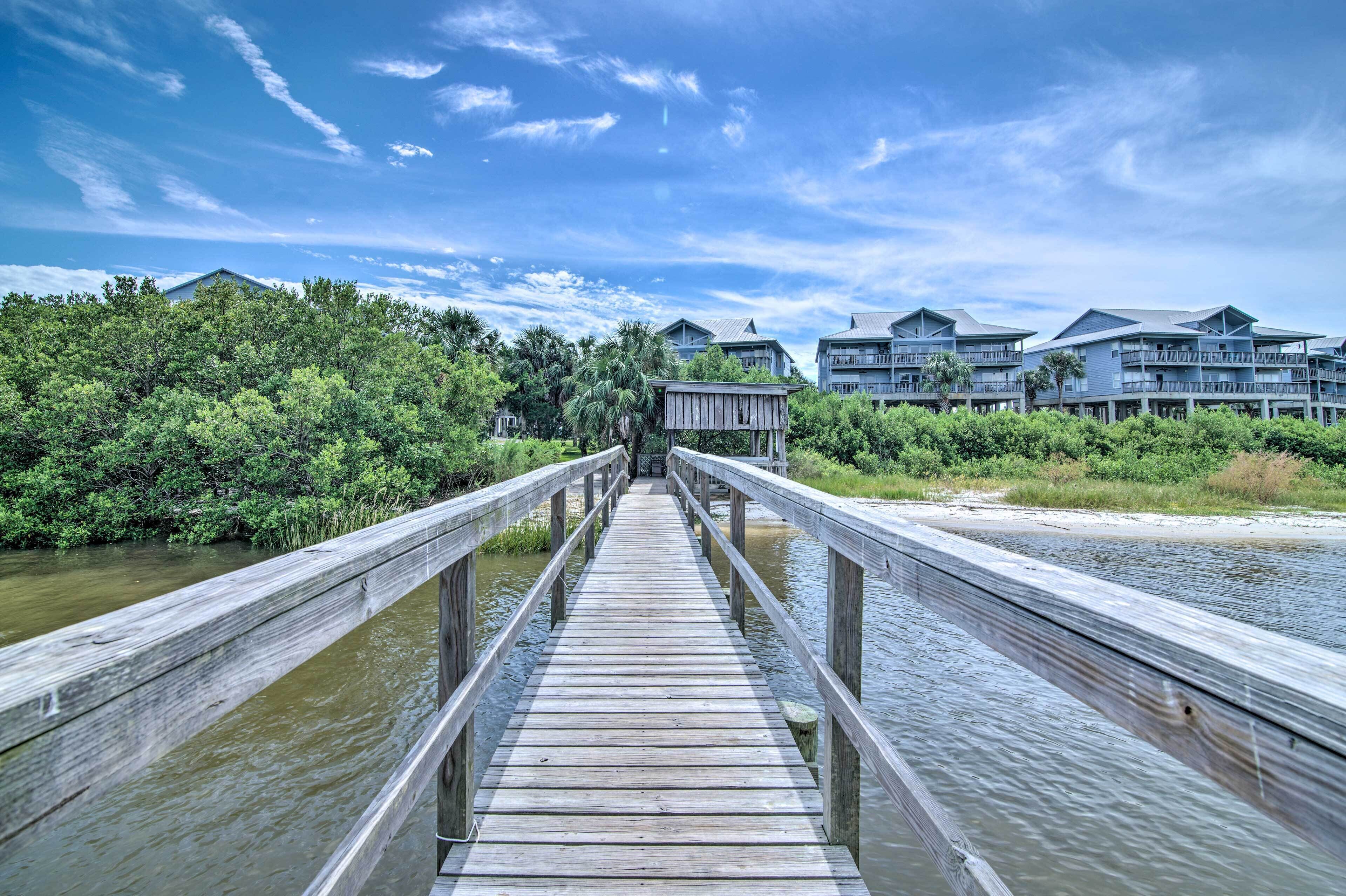 A wooden pier leading to a building with multiple balconies, surrounded by lush greenery and a clear sky.