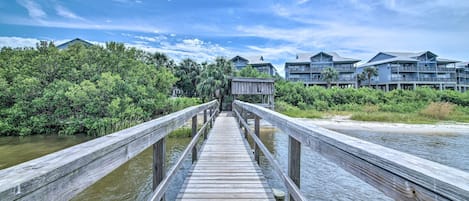 A wooden pier leading to a building with multiple balconies, surrounded by lush greenery and a clear sky.