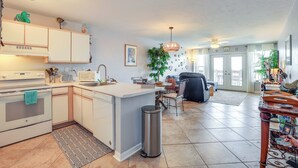 Een moderne keuken met witte kasten, een ingebouwde magnetron en een klein eiland. Er is een zithoek met een zwarte stoel en een tafel, en een groot raam met gordijnen.