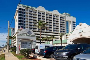 A multi-story hotel with a sign indicating it is a hotel and suites. There are several parked cars in front, and palm trees are visible. - Beachfront Resort Condo, Book for Bike Week (Daytona Beach)