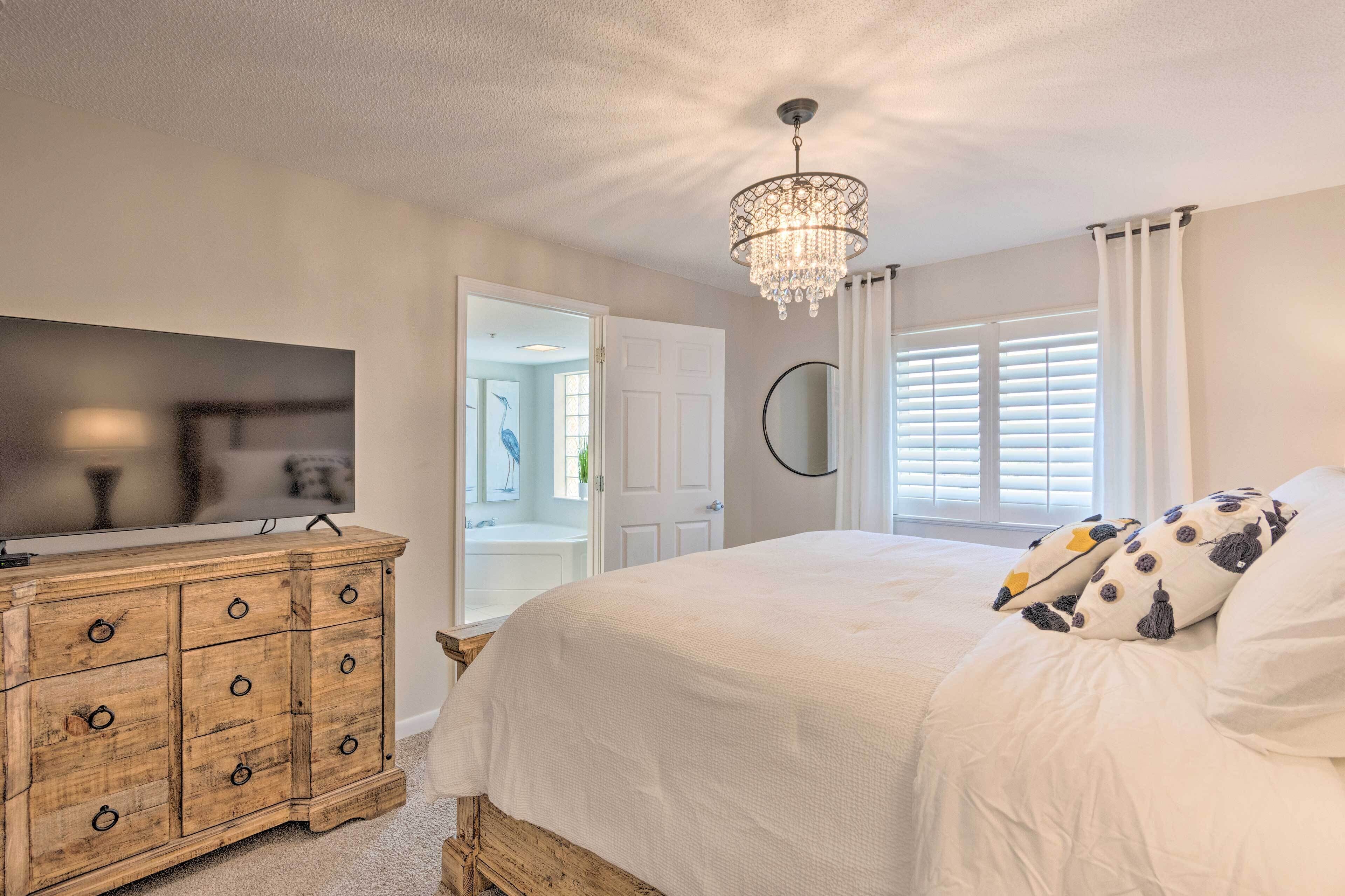 A bedroom with a wooden dresser, a bed with white bedding, a chandelier, and a window with blinds.